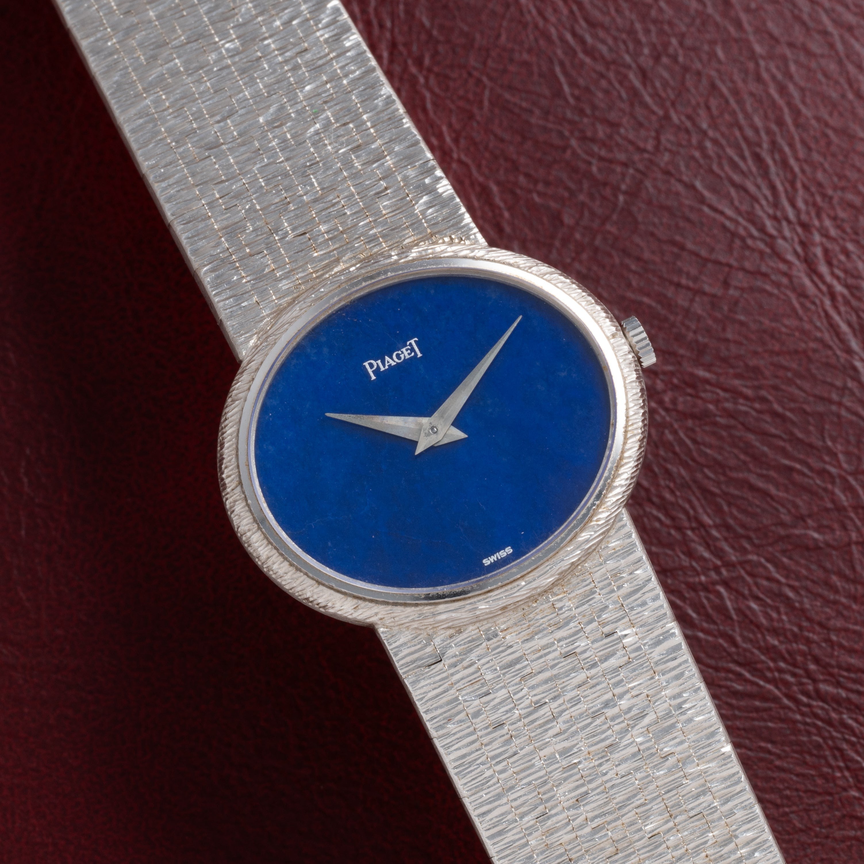 A Piaget White Gold Lapis Lazuli Dress Watch with a blue face rests on a dark red leather surface.