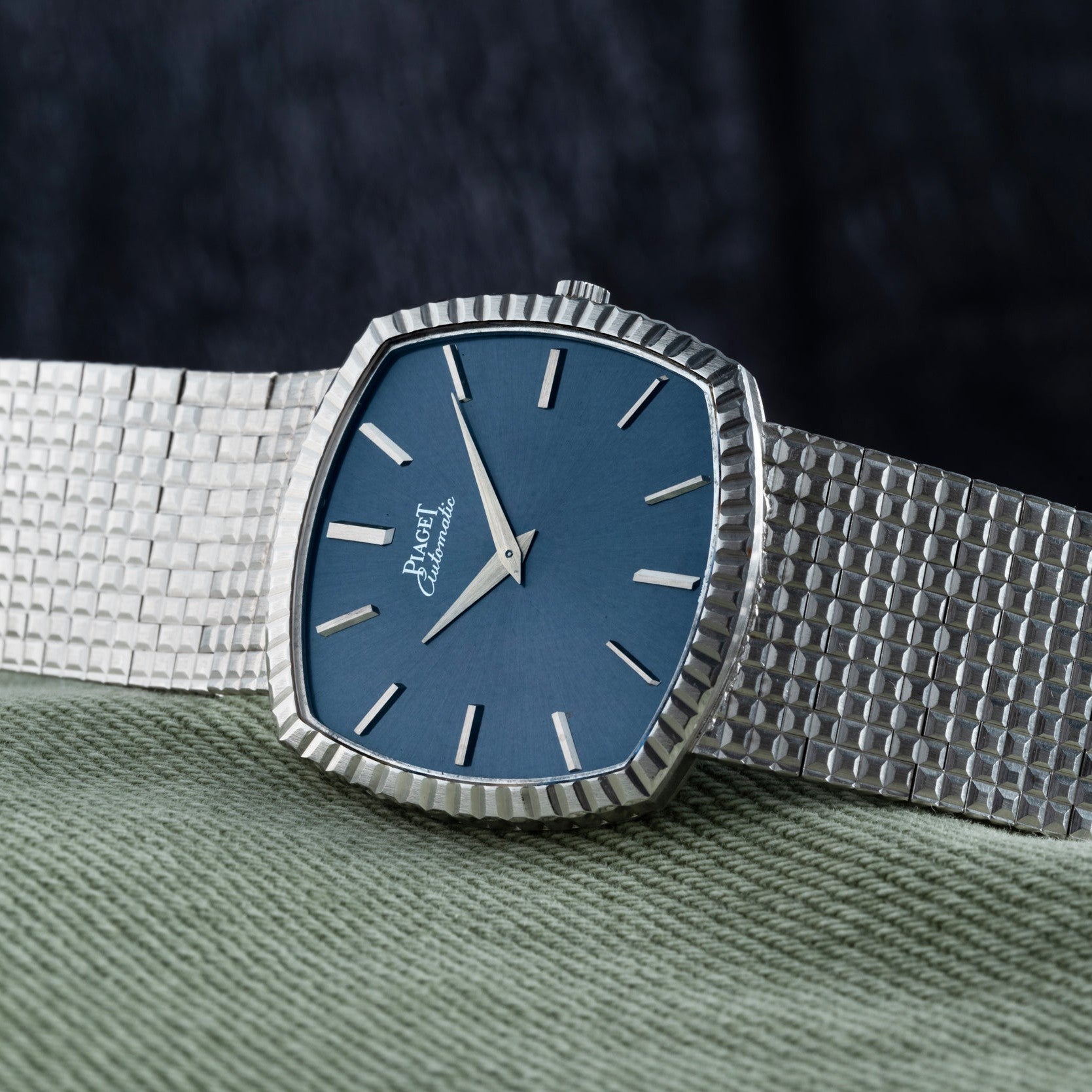 The Piaget Sqound White Gold Dress Watch features a silver band and blue dial with silver hour markers, displayed on green fabric.