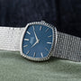 The Piaget Sqound White Gold Dress Watch features a silver band and blue dial with silver hour markers, displayed on green fabric.