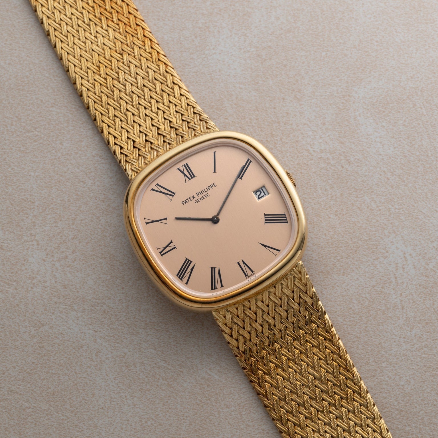 A Patek Philippe Ellipse Jumbo gold wristwatch with Roman numerals and a mesh band rests on a light textured surface.