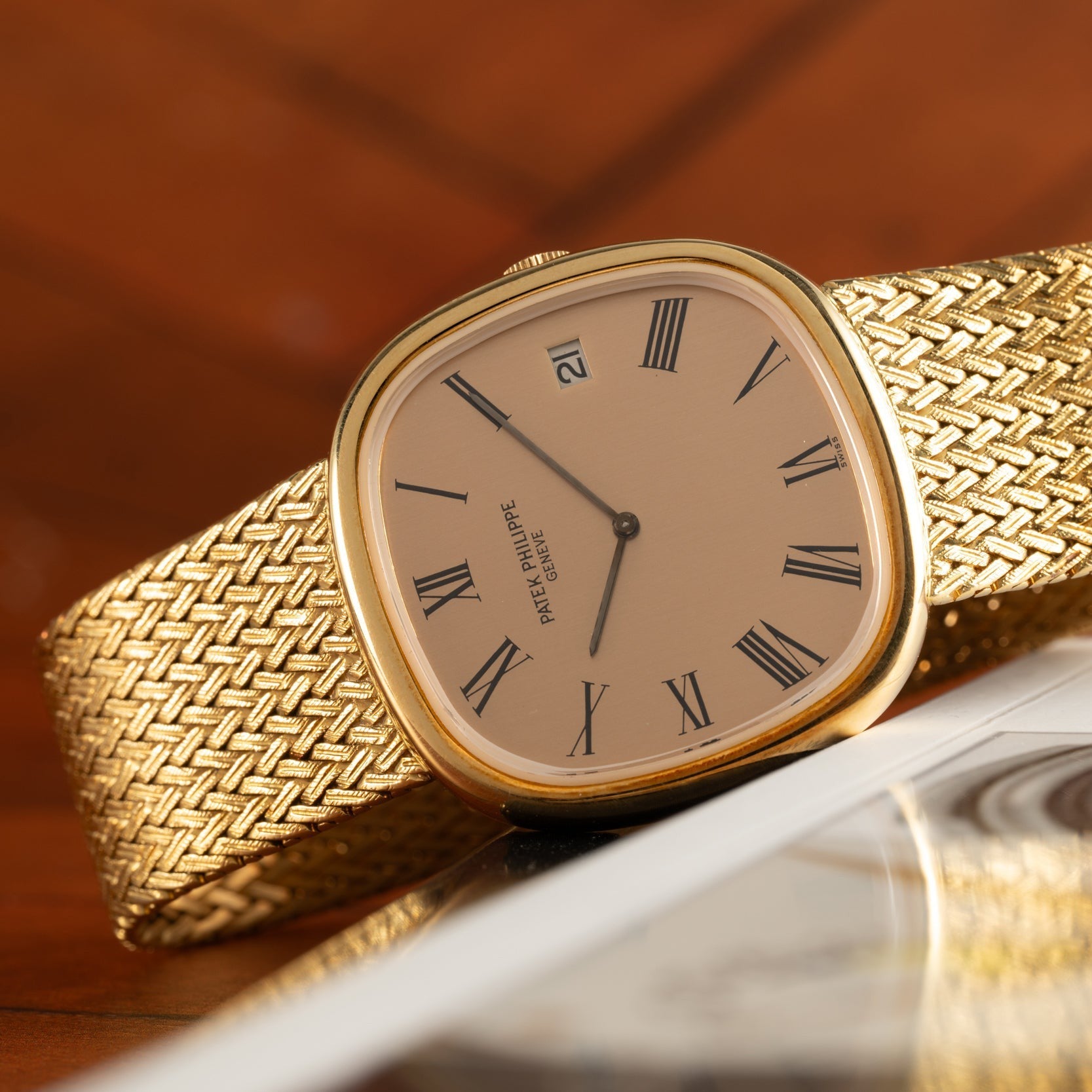 A Patek Philippe Ellipse Jumbo gold watch with a woven band and Roman numerals, resting on a wooden surface.