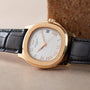 A Patek Philippe Aquanaut gold wristwatch with a white dial, Roman numerals, and a black leather strap rests on a cork surface.
