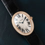 The Cartier Baignoire Jumbo watch features a diamond bezel, black Roman numerals, and a black leather strap, displayed on a dark surface.
