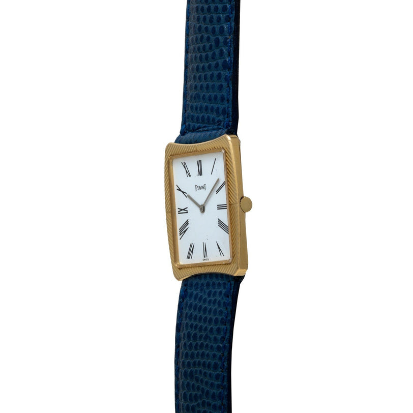 The Cartier Protocole is a gold rectangular watch featuring a white face, Roman numerals, and a dark blue textured leather strap.