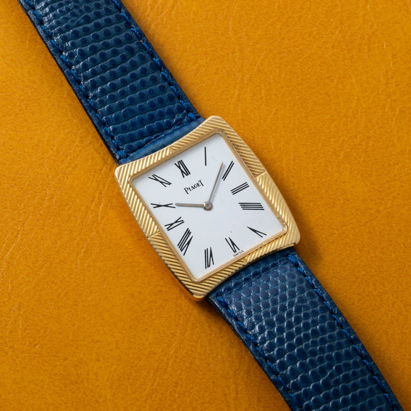 A Cartier Protocole gold wristwatch with a blue textured strap rests on a yellow surface.