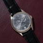 A Patek Philippe Annual Calendar Moonphase wristwatch with a black leather strap rests on a dark red textured surface.
