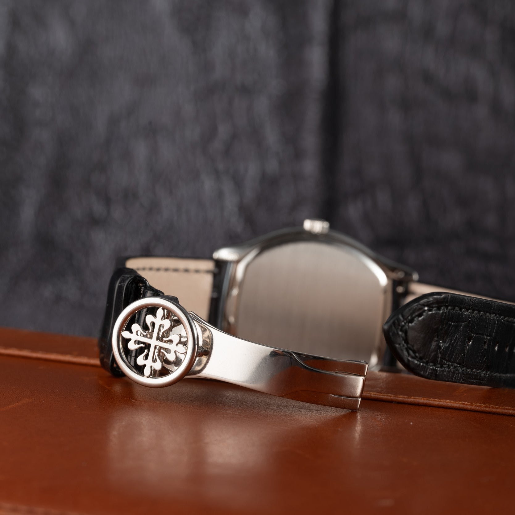 The Patek Philippe Tonneau Perpetual Calendar watch features a black leather strap and silver clasp, elegantly displayed on a brown surface with a blurred background.