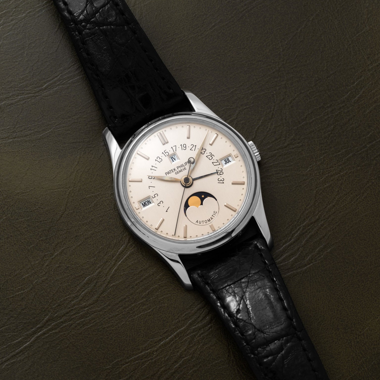 Patek Philippe Retrograde Perpetual Calendar: silver wristwatch with black leather strap, moon phase display, and date indicators on a dark leather surface.