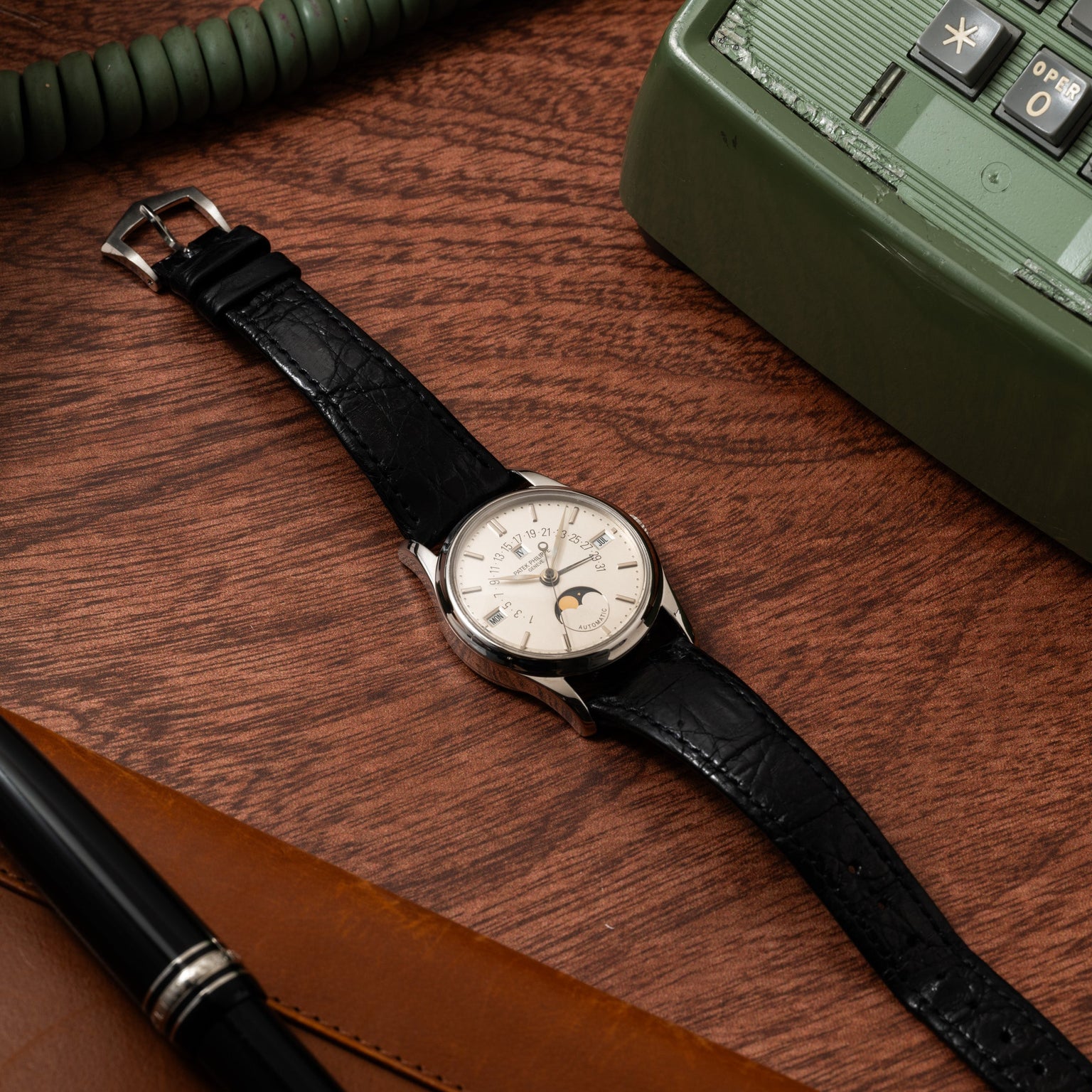 A Patek Philippe Retrograde Perpetual Calendar wristwatch with a black leather strap rests on a wooden desk beside a green phone and a brown leather accessory.
