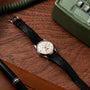 A Patek Philippe Retrograde Perpetual Calendar wristwatch with a black leather strap rests on a wooden desk beside a green phone and a brown leather accessory.