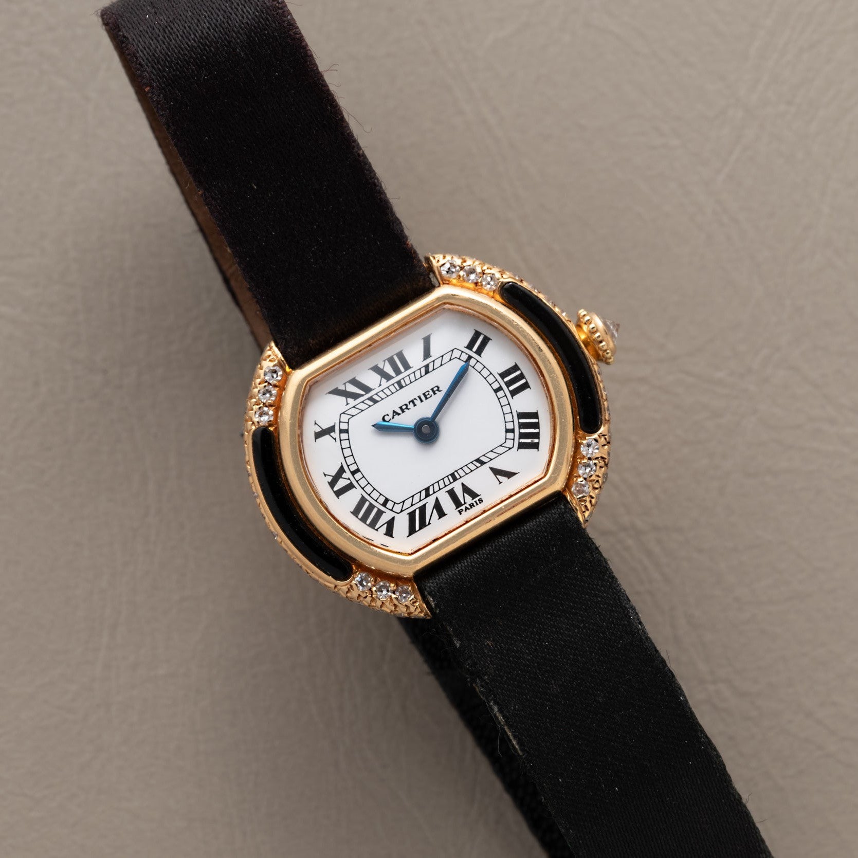 Cartier Ellipse watch featuring a black strap, gold case, Roman numerals, and diamond accents displayed on a beige surface.