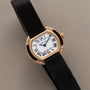 Cartier Ellipse watch featuring a black strap, gold case, Roman numerals, and diamond accents displayed on a beige surface.