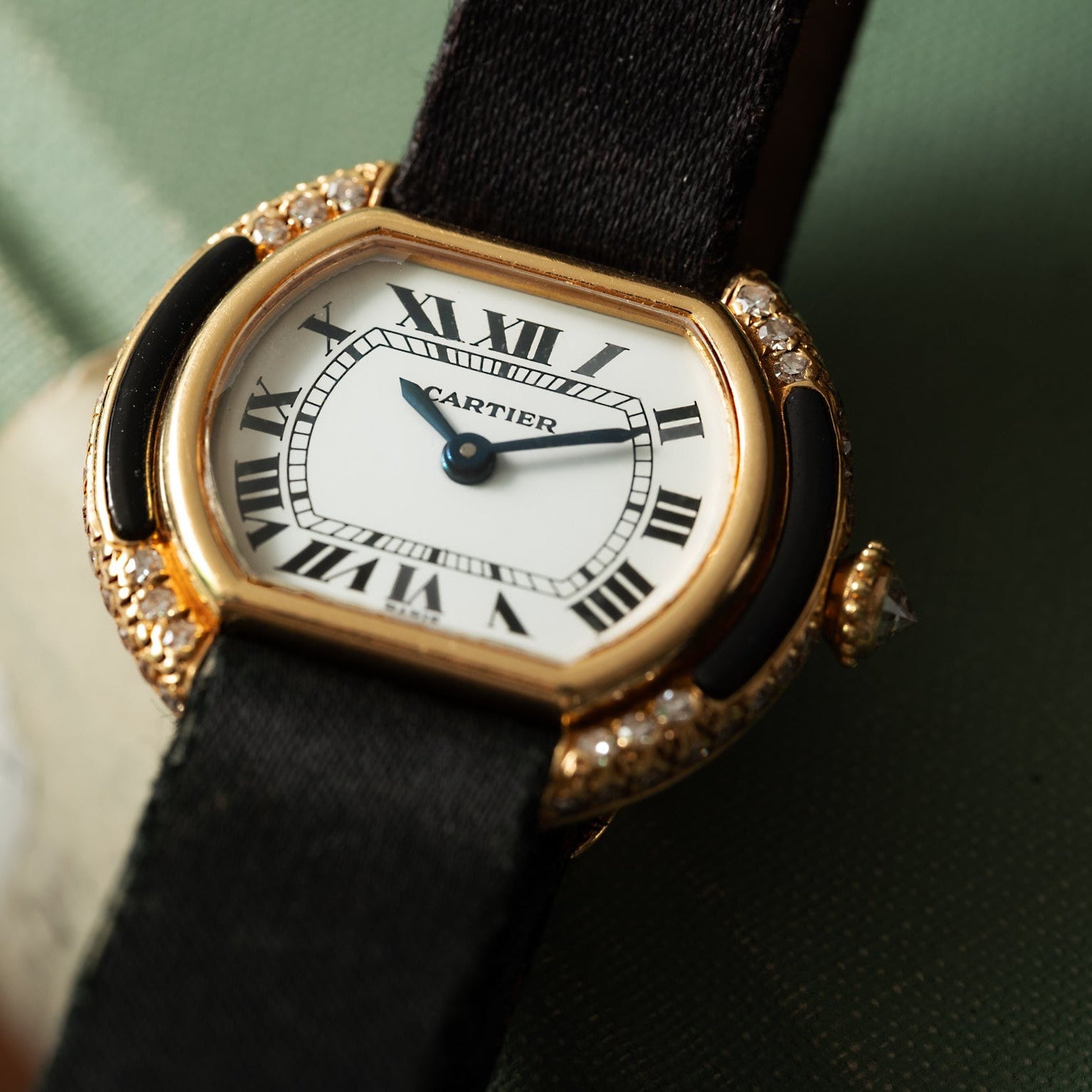 Cartier Ellipse gold wristwatch with a black strap, Roman numerals, and diamond accents displayed on a green surface.