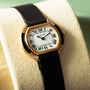 The Cartier Ellipse watch features a black strap and Roman numeral face, elegantly displayed on a beige cushion.
