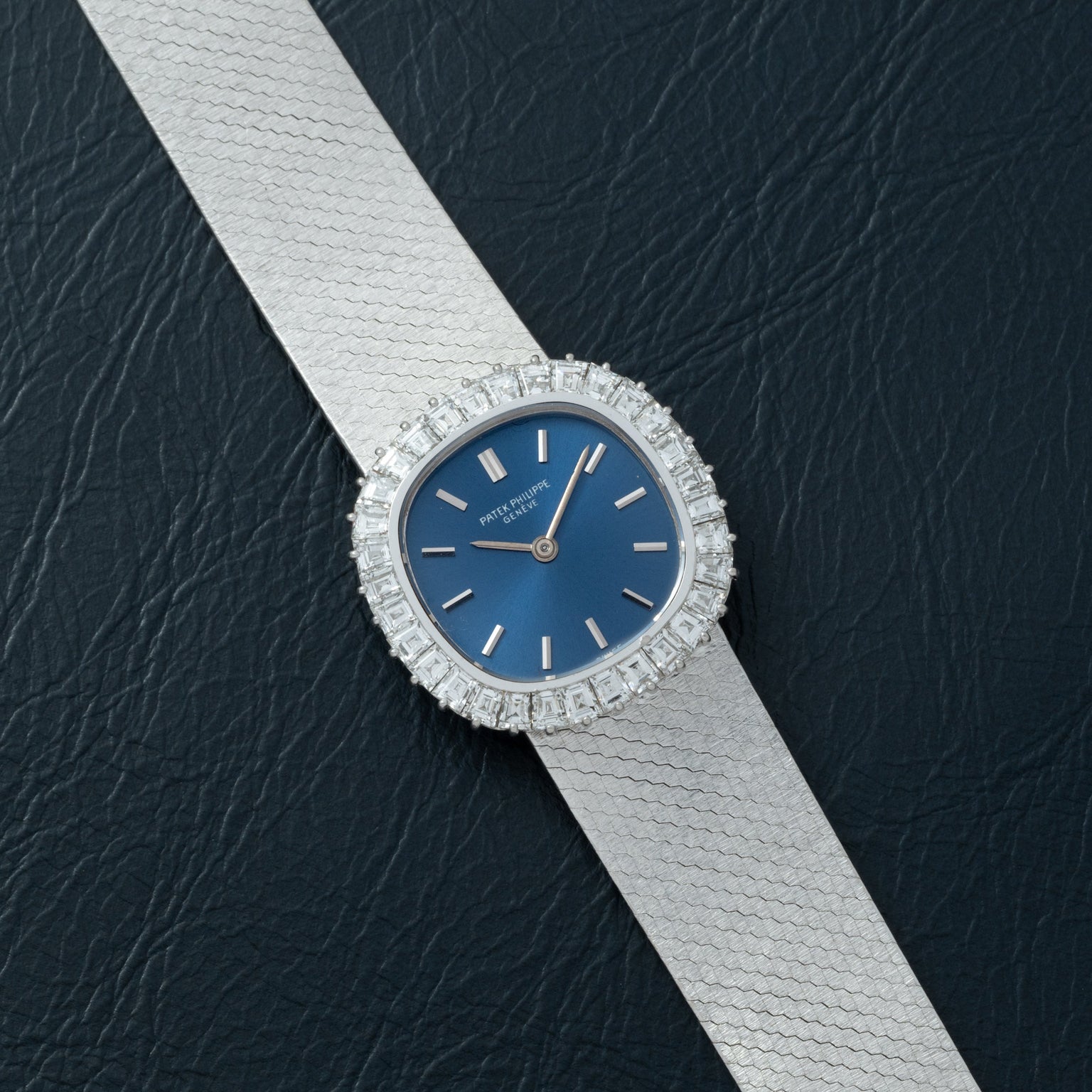 The Patek Philippe Ellipse Ladies Dress Watch features a silver case, blue face, and diamond bezel, elegantly displayed on black textured leather.