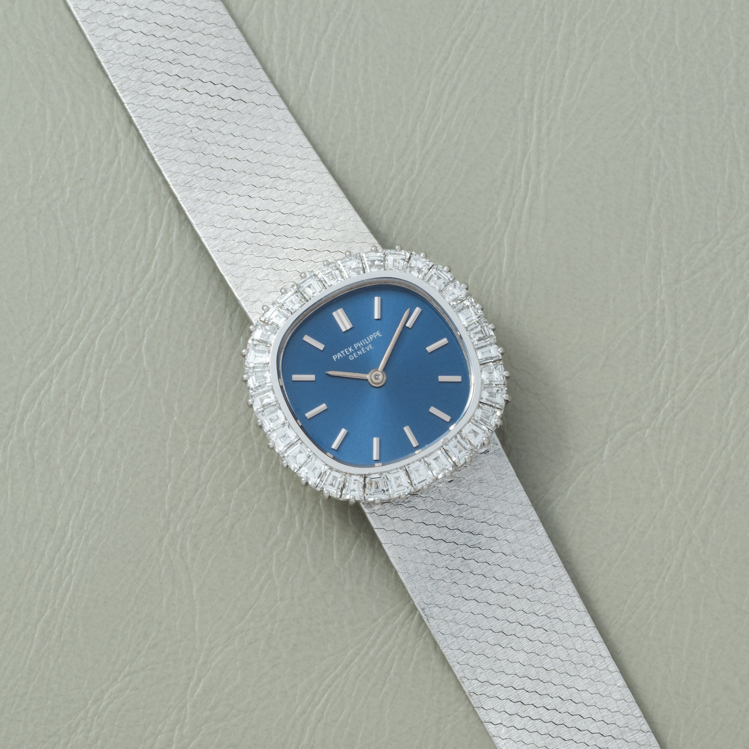 The Patek Philippe Ellipse Ladies Dress Watch features a blue face, diamond bezel, and silver strap, displayed on a light gray textured surface.