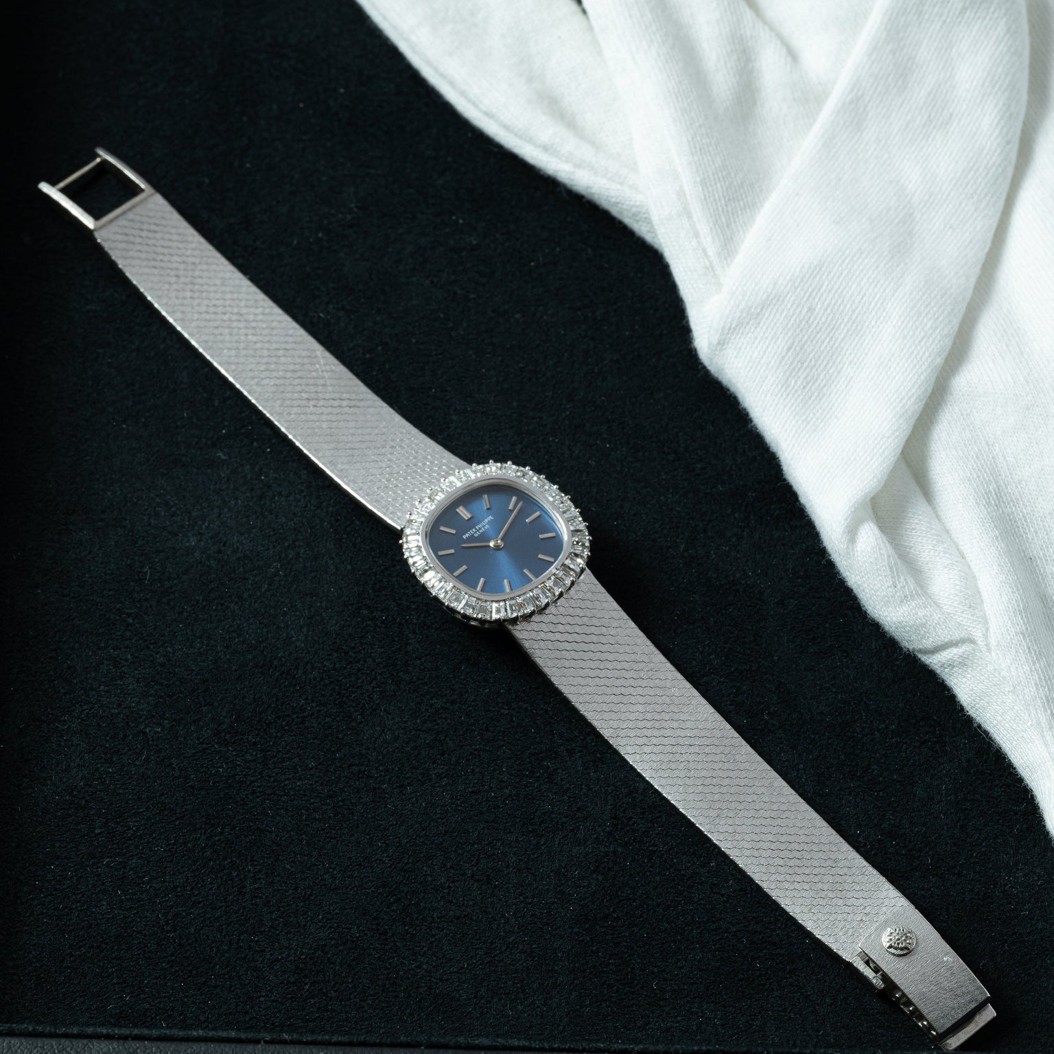 The Patek Philippe Ellipse Ladies Dress Watch, featuring a blue face and diamond bezel, is displayed on a black surface beside a white cloth.