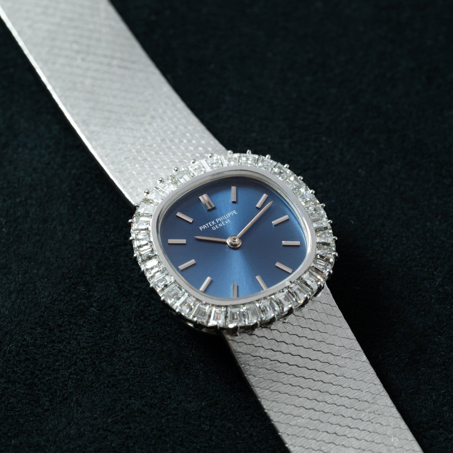 A Patek Philippe Ellipse Ladies Dress Watch in silver with a blue face and diamond bezel is displayed against a textured dark background.