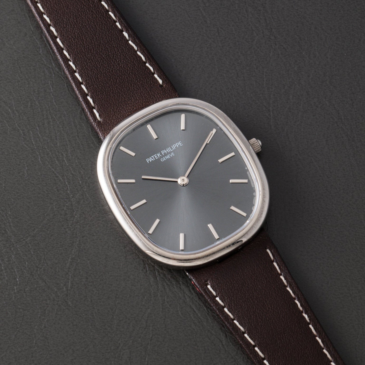 A Patek Philippe Ellipse watch with a silver case, gray face, and brown leather strap displayed on a black surface.