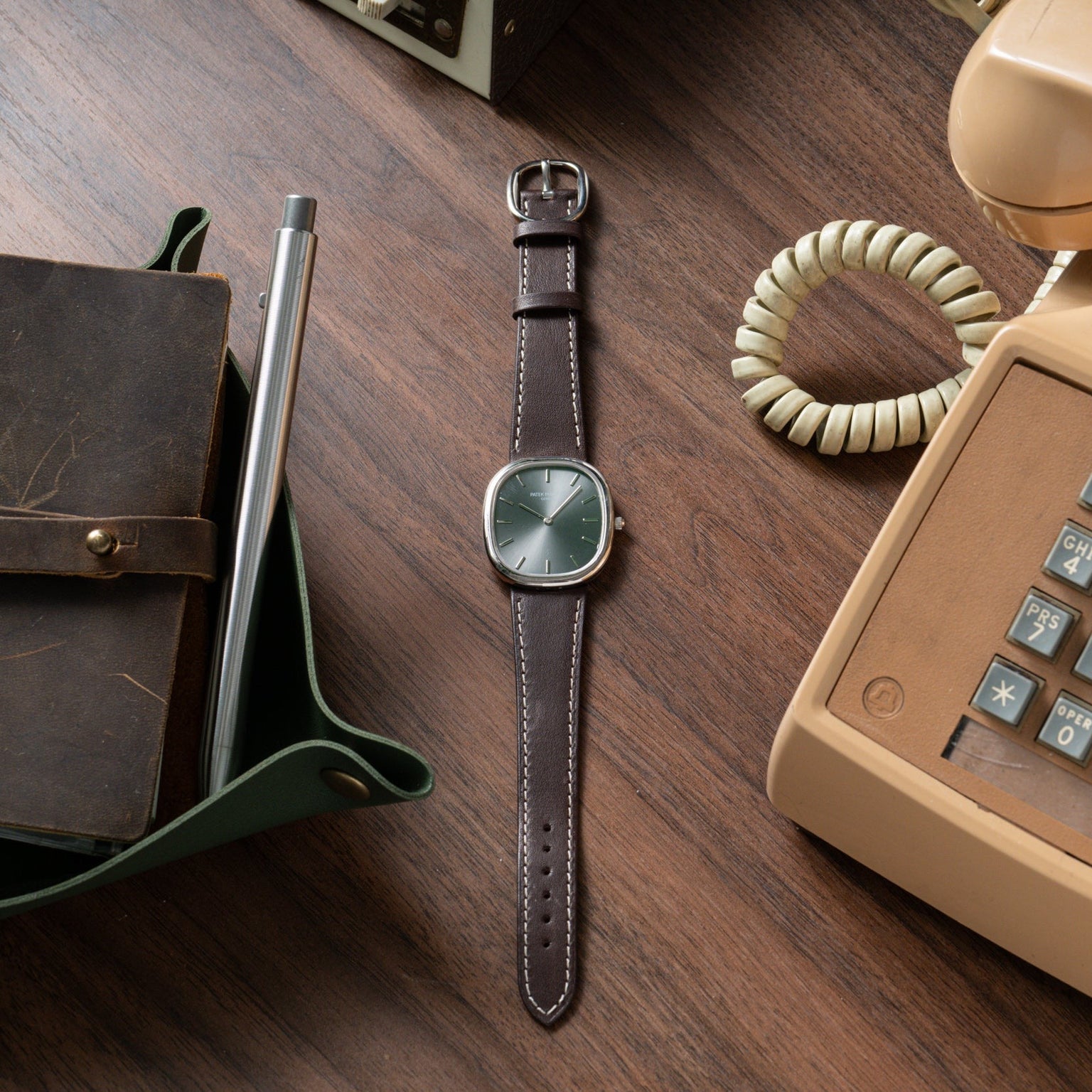 A Patek Philippe Ellipse brown leather wristwatch rests on a wooden desk beside a rotary phone, notebook, and pen.