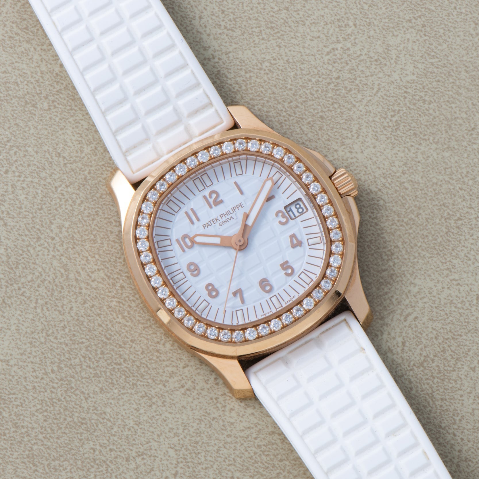 A Patek Philippe Aquanaut Ladies gold watch with a diamond bezel and white strap rests on a light textured surface.