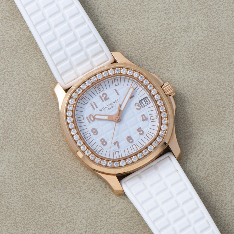 A Patek Philippe Aquanaut Ladies gold watch with a diamond bezel and white strap rests on a light textured surface.