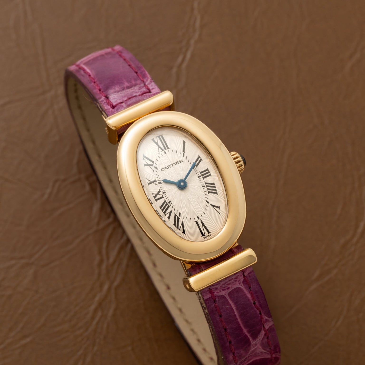 Cartier Baignoire watch with gold oval case, roman numerals, and a purple leather strap placed on a brown textured surface.
