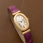 Cartier Baignoire watch with gold oval case, roman numerals, and a purple leather strap placed on a brown textured surface.