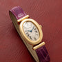 Cartier Baignoire watch with a gold oval case, Roman numerals, and a purple leather strap displayed on a red textured surface.