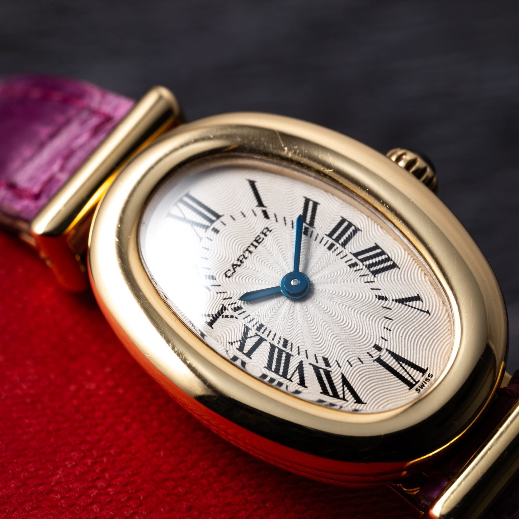 The Cartier Baignoire watch features a gold oval case, Roman numerals, blue hands, and a purple strap, displayed on a red and black surface.
