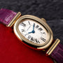 The Cartier Baignoire watch features a gold oval case, Roman numerals, and a purple leather strap, displayed on a red surface.