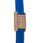 The Cartier Tank Louis Diamonds watch features a gold case, blue textured strap, and diamond and pink gemstone face and bezel, displayed from an angle.