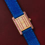The Cartier Tank Louis Diamonds watch features a jeweled face, gold case, blue strap, and pink accents, elegantly displayed on burgundy leather.