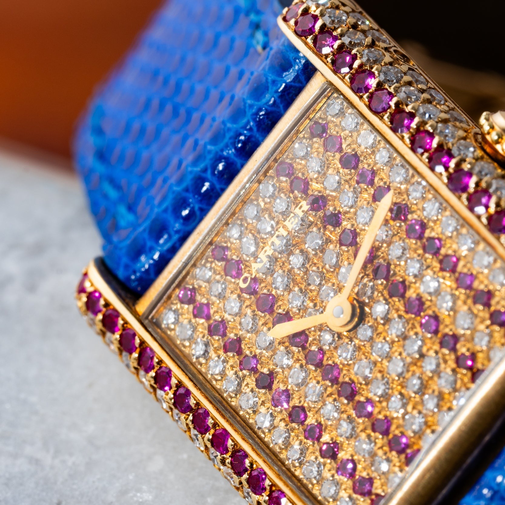 Close-up of the Cartier Tank Louis Diamonds watch featuring a blue strap, gold case, and a diamond and ruby-encrusted face.