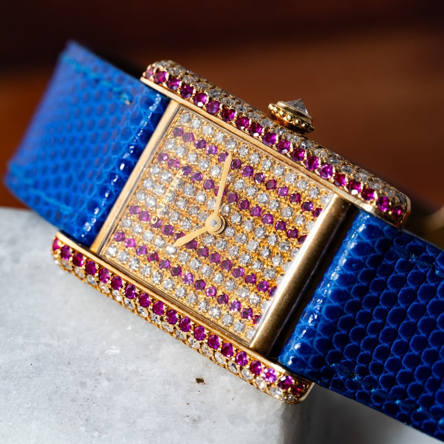 The Cartier Tank Louis Diamonds watch features a gem-studded face and bezel, a blue textured strap, and is elegantly displayed on a marble surface.
