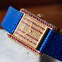 The Cartier Tank Louis Diamonds watch features a gem-studded face and bezel, a blue textured strap, and is elegantly displayed on a marble surface.