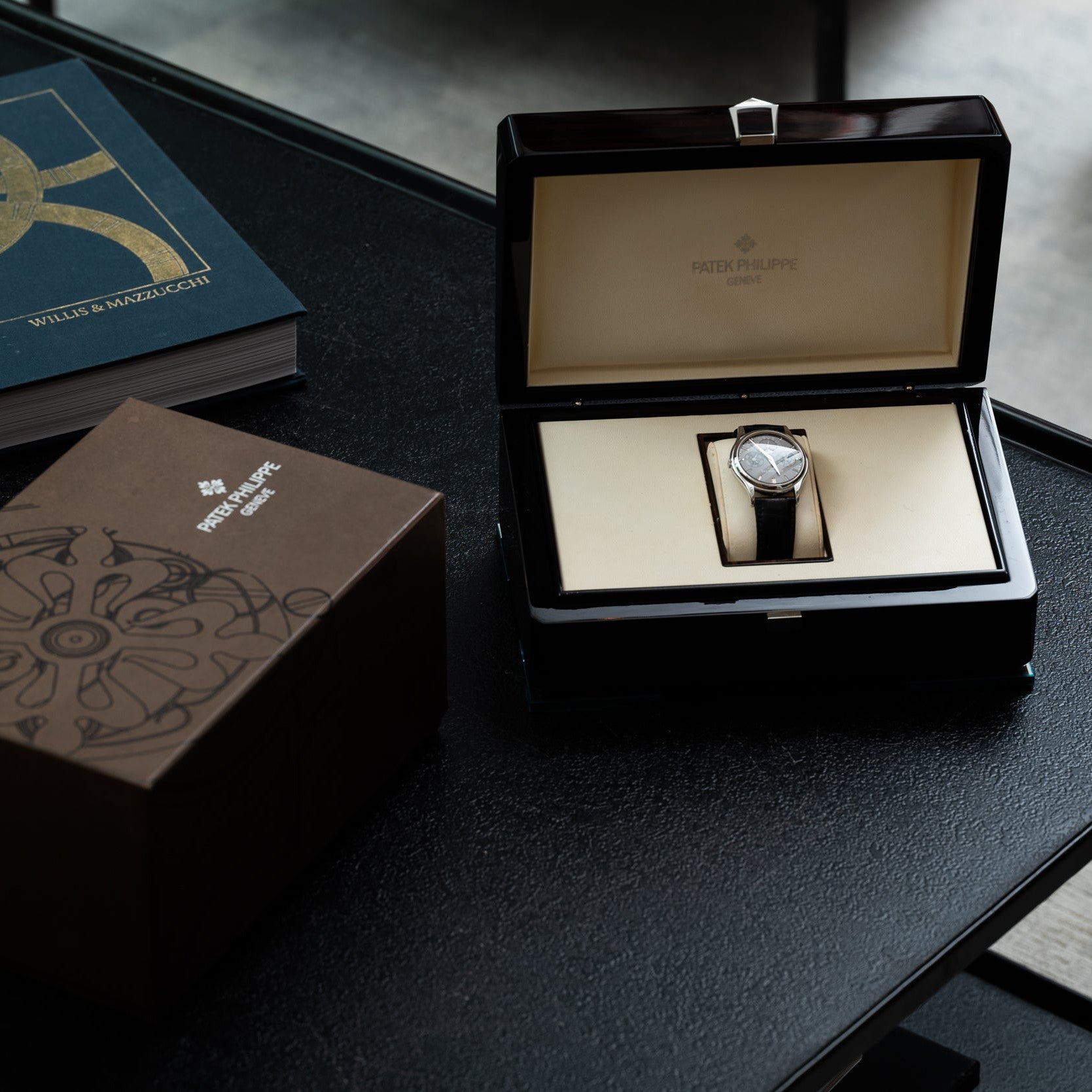 A Patek Philippe Perpetual Calendar watch displayed in an open box with its packaging and a book on a dark table.