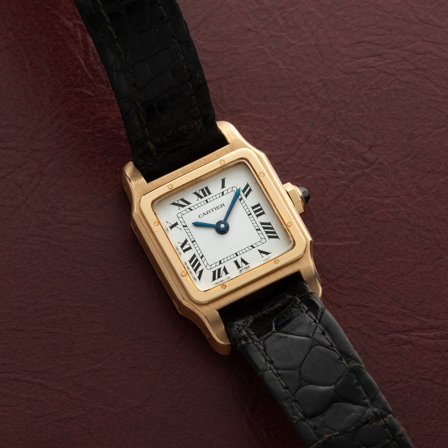 The Cartier Santos Mini watch with a black leather strap rests on a textured dark red surface.