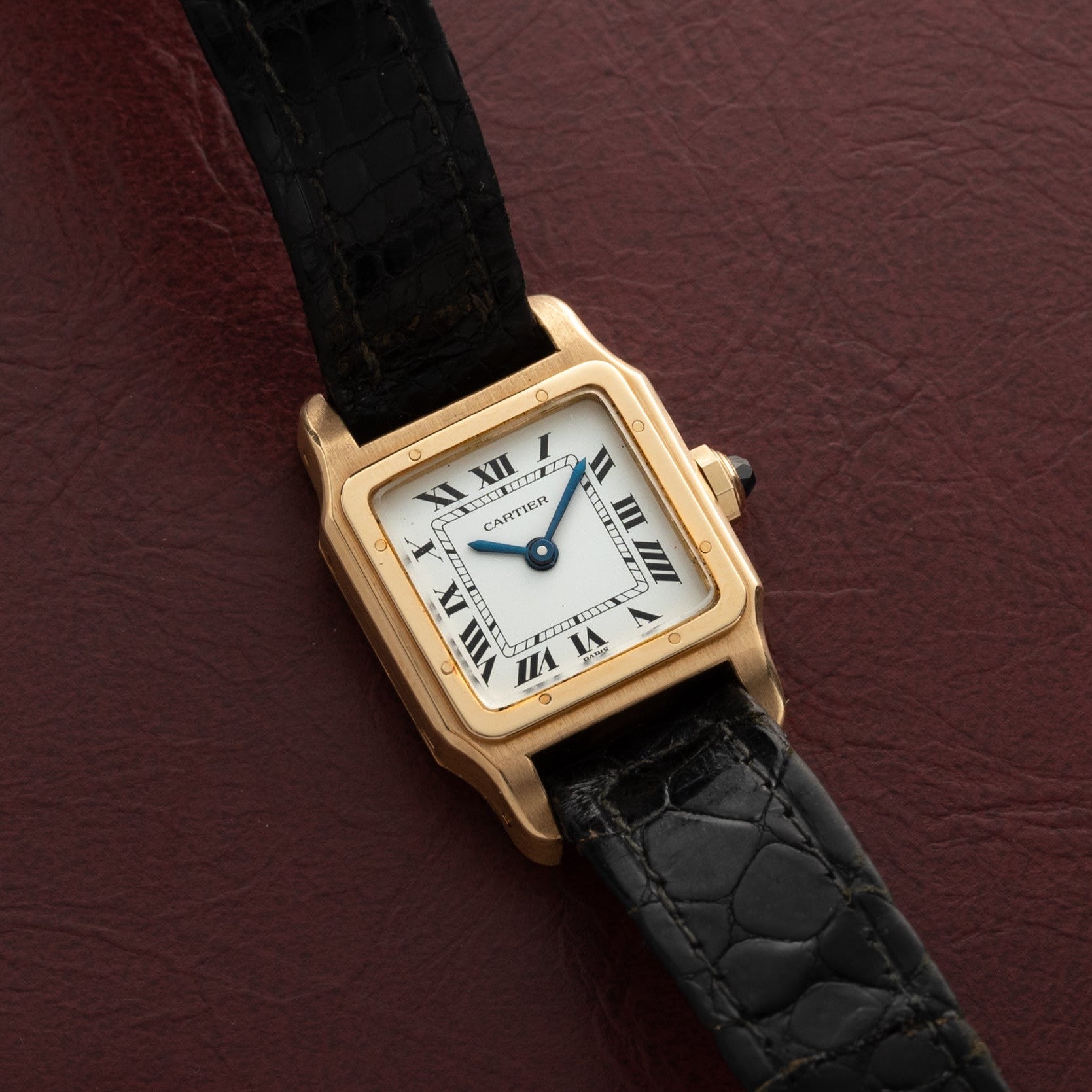 The Cartier Santos Mini watch with a black leather strap rests on a textured dark red surface.