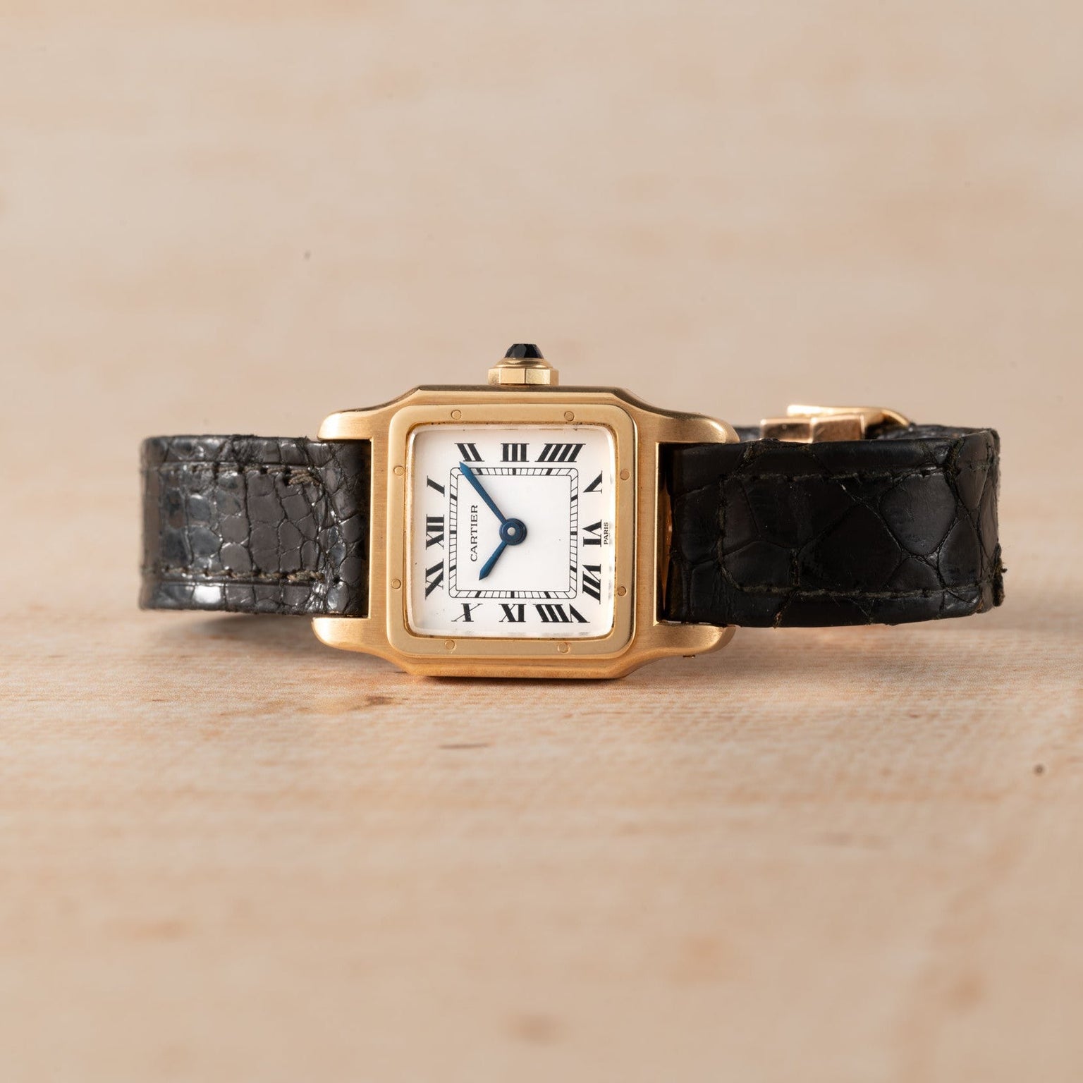 Cartier Santos Mini gold square wristwatch with Roman numerals and black leather strap displayed on a light wooden surface.