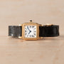 Cartier Santos Mini gold square wristwatch with Roman numerals and black leather strap displayed on a light wooden surface.