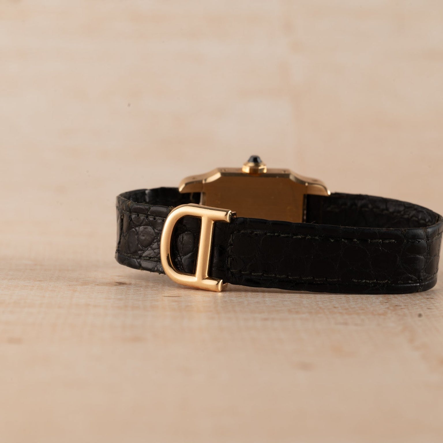 A Cartier Santos Mini black leather watch with a gold buckle and rectangular gold case is displayed on a light wooden surface.