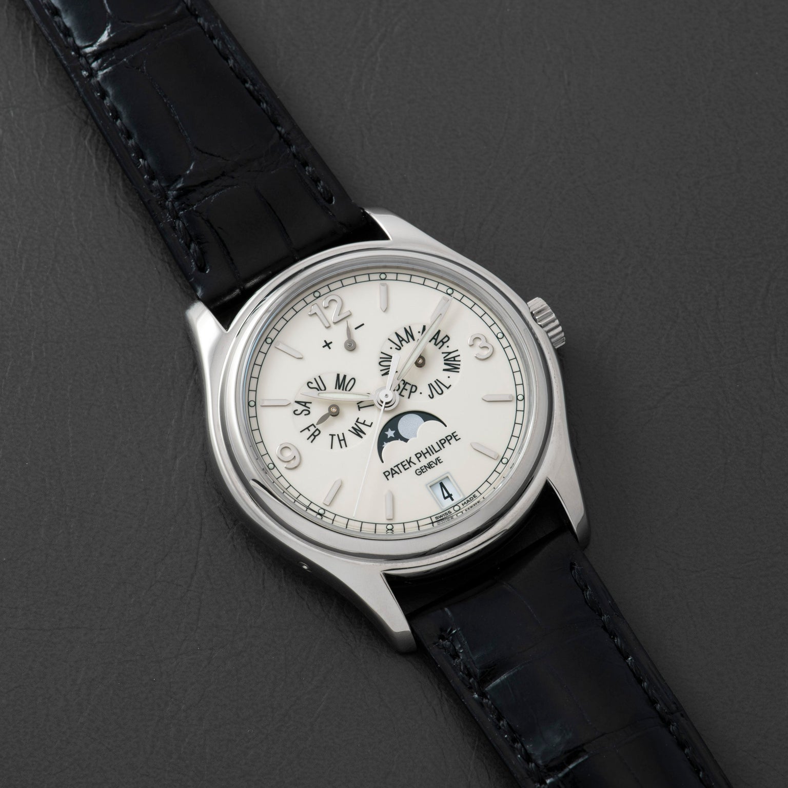 The Patek Philippe Annual Calendar Moonphase, a white gold luxury watch with a white dial, black leather strap, Roman numerals, moonphase display, and date window, is displayed on a dark textured surface.