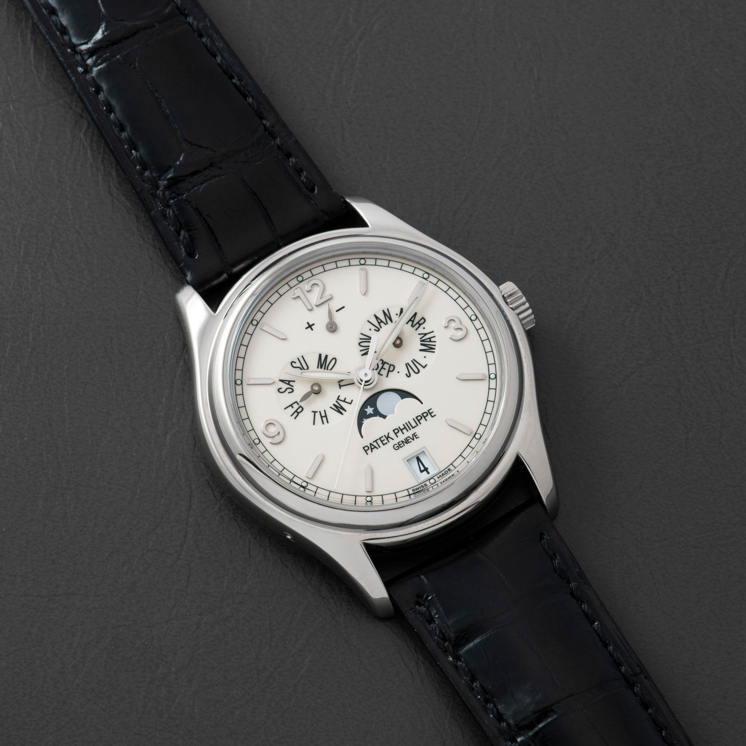 The Patek Philippe Annual Calendar Moonphase, a white gold luxury watch with a white dial, black leather strap, Roman numerals, moonphase display, and date window, is displayed on a dark textured surface.