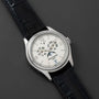 The Patek Philippe Annual Calendar Moonphase, a white gold luxury watch with a white dial, black leather strap, Roman numerals, moonphase display, and date window, is displayed on a dark textured surface.