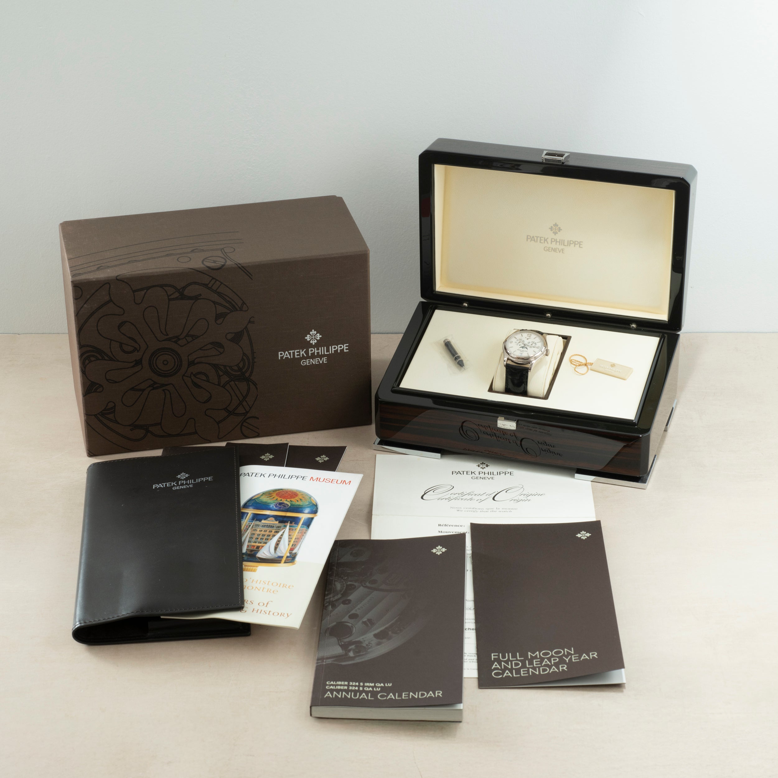 A Patek Philippe Annual Calendar Moonphase watch in white gold, presented with a brown box, wooden case, pen, key, black booklet holder, and several instruction and certificate booklets on a light surface.
