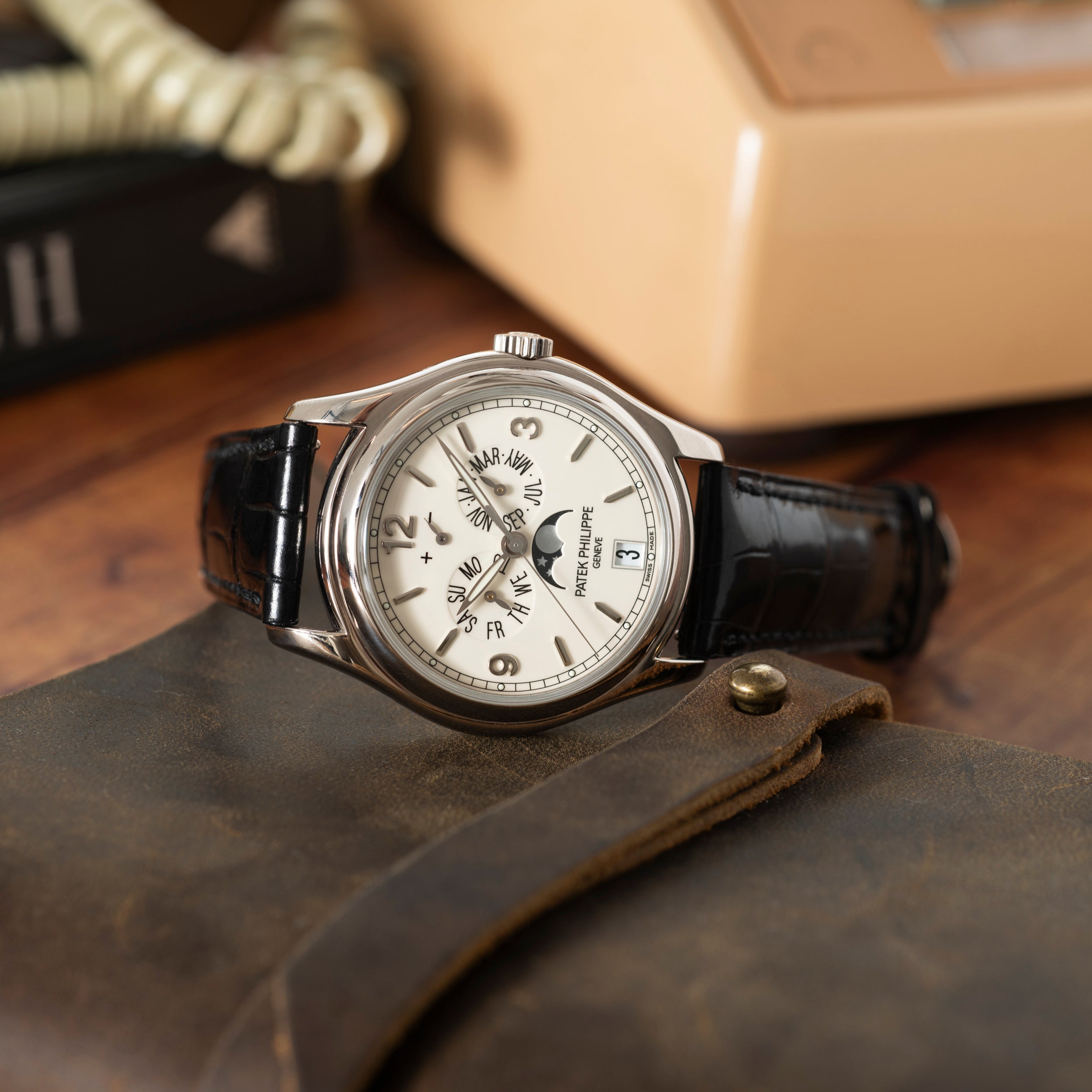A Patek Philippe Annual Calendar Moonphase wristwatch with a black leather strap rests on a brown case, while an old beige telephone and a book are blurred in the background. The watch features an annual calendar, day, date, and 24-hour indicators.