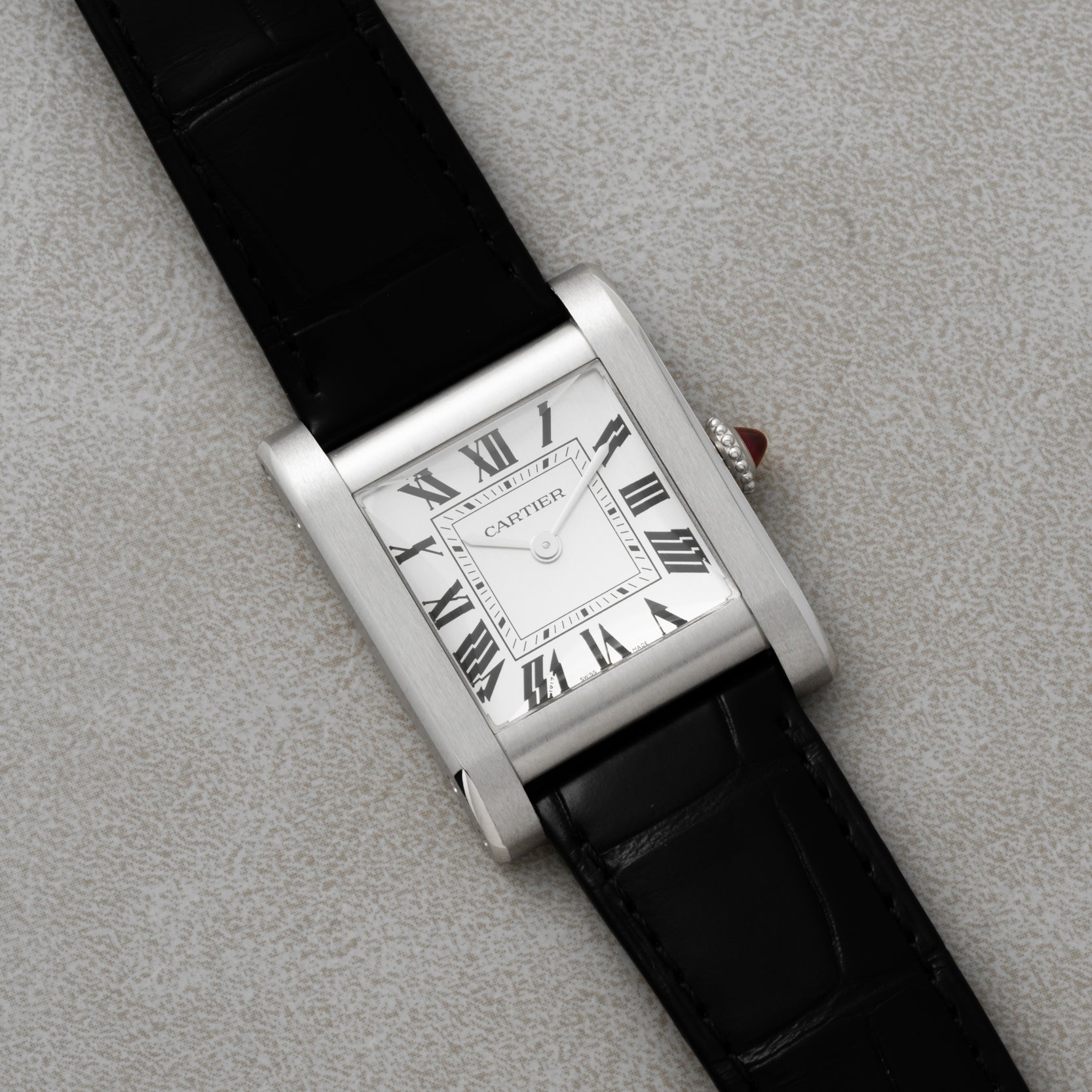 The Cartier Tank Normale watch features a rectangular case, black leather strap, and Roman numeral dial, displayed on a light gray surface.