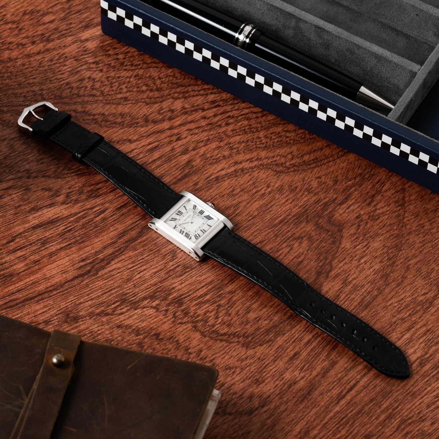 The Cartier Tank Normale, featuring a black leather strap and square face, rests on a wooden surface beside a pen box and notebook.