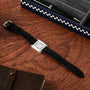 The Cartier Tank Normale, featuring a black leather strap and square face, rests on a wooden surface beside a pen box and notebook.